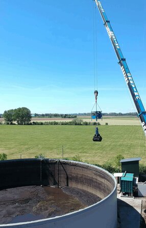 Ein Mitarbeiter sitzt mit Schutzanzug, Gasmaske & Handschuhen auf einem Mini-Bagger der vorne einen Aufsatz zum zusammenschieben von Materalien hat. Er arbeitet im inneren eines Tanks und schieb eine Schlamm ähnliche Substanz rum.