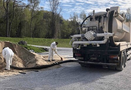 Zu sehen ist ein LKW mit Tank von hinten, an dem ein Schlauch befestigt ist, welcher von einem Arbeiter genutzt wird um eine Sand ähnlichen Berg aufzusaugen. Ein anderer Arbeiter Kehr den Sand der neben dem Berg zusammen.
