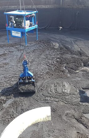 Ansicht von oben, Vogelperspektive, eine Tanks mit schlammartigem Inhalt, welcher durch einen kleinen gelben Bagger, rechts im Bild, zusammen geschoben wird. Ein königsblauer Greifarm, in der Mitte des Bildes, heb beziehungsweise lädt die schlammartige Masse mit seinem Greifer auf..