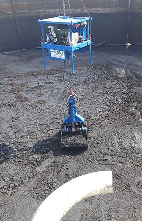 Ansicht von oben, Vogelperspektive, eine Tanks mit schlammartigem Inhalt, welcher durch einen kleinen gelben Bagger, rechts im Bild, zusammen geschoben wird. Ein königsblauer Greifarm, in der Mitte des Bildes, heb beziehungsweise lädt die schlammartige Masse mit seinem Greifer auf..