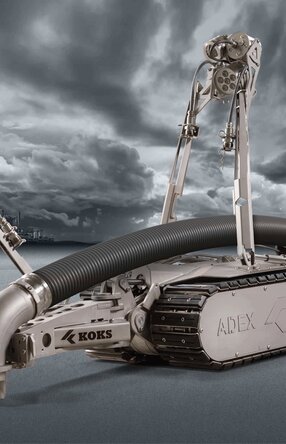 Roboter mit Schlauch, der Richtung Boden zeigt, und Kameras, zur Steuerung durch einen Mitarbeiter. Im Hintergrund ist weit in der Ferne Industrie-Landschaft zu sehen, zusammen mit dunkelgrauen Wolken und Boden.