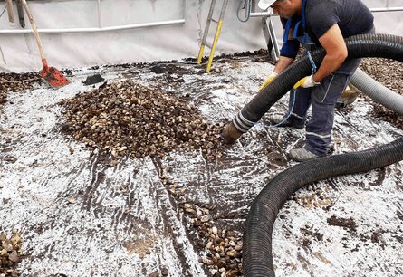 Zu sehen ist ein Arbeiter in Arbeitskleidung und mit Schutzhelm, der mit einem Rohr zusammengeschobenes Kies von einen Dach aufsaugt.