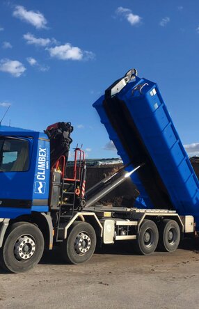 Zu sehen ist ein LKW, in der Mitte des Bildes, mit gekippter Ladefläche wie er Dreck ablädt. Hinter dem LKW steht eine Halle vor der weiterer Dreck liegt.