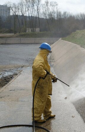 Es steh eine Person mit gelbem Ganzkörperanzug und königsblauen Arbeitshelm in einer offenen, Kanal ähnlichen, Grube und hält dabei einen Hochdruckstrahler auf die Innenwand.