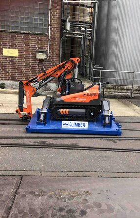 Roboter, orangefarben, mit gesenktem Greifarm stehend auf einer königsblauen Platte die über ein Schienensystem auf einem Betriebshof fährt. Im Hintergrund sind eine Backstein-Fassade und Silos oder Tanks zu sehen.