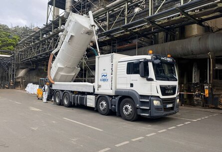 Weißer LKW mit Climbex Logos und einem gekippten Tank hinten drauf steht vor einem Fabrikgebäude, außen verlaufen Metallstützen. Hinten links neben dem LKW steht ein Mitarbeiter in weißem Anzug und mit orangefarbenen Arbeitshelm, er steuert den Tank. Hinter dem LKW und Mitarbeiter ist ein Gabelstapler zu sehen.
