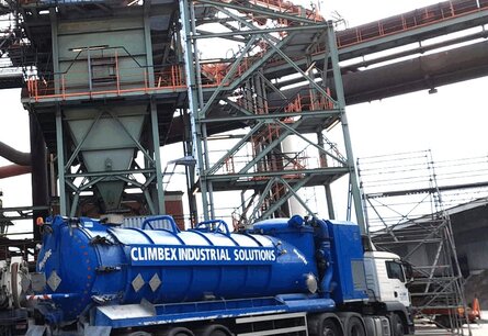 Zu sehen ist das Äußere eines Fabrikgeländes mit diversen Rohren und Metallgeländern, außerdem steht darin eine trichterförmige Anlage. Vor der Anlage steht ein LKW mit einem großen Tank.